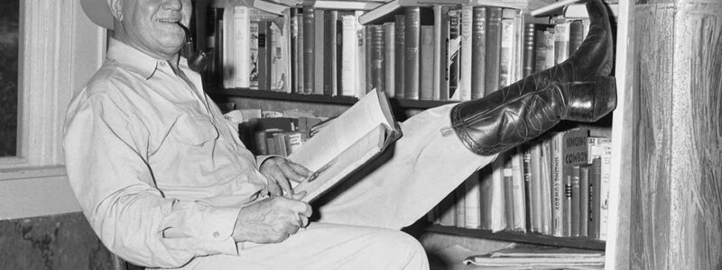 Photo of J. Frank Dobie in sitting in front of bookshelf with cowboy-booted foot raised high and resting on a shelf