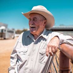 An older cowboy leaning on a fence and smiling