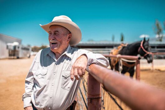 An older cowboy leaning on a fence and smiling